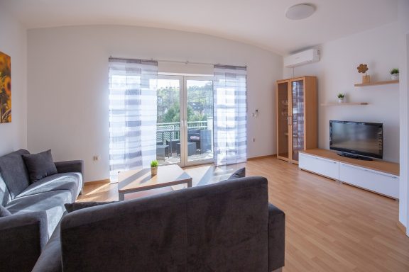 Wohnbereich Heller Wohnraum mit grauem Sofa, Glastisch, TV und Balkonzugang.